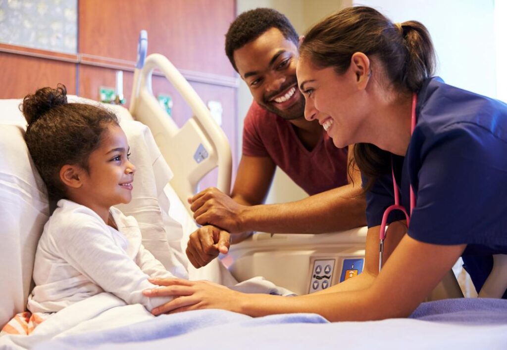 Nurse in dark blue scrubs using a pink stethoscope on a smiling girl in a hospital bed, with her father sitting beside her.